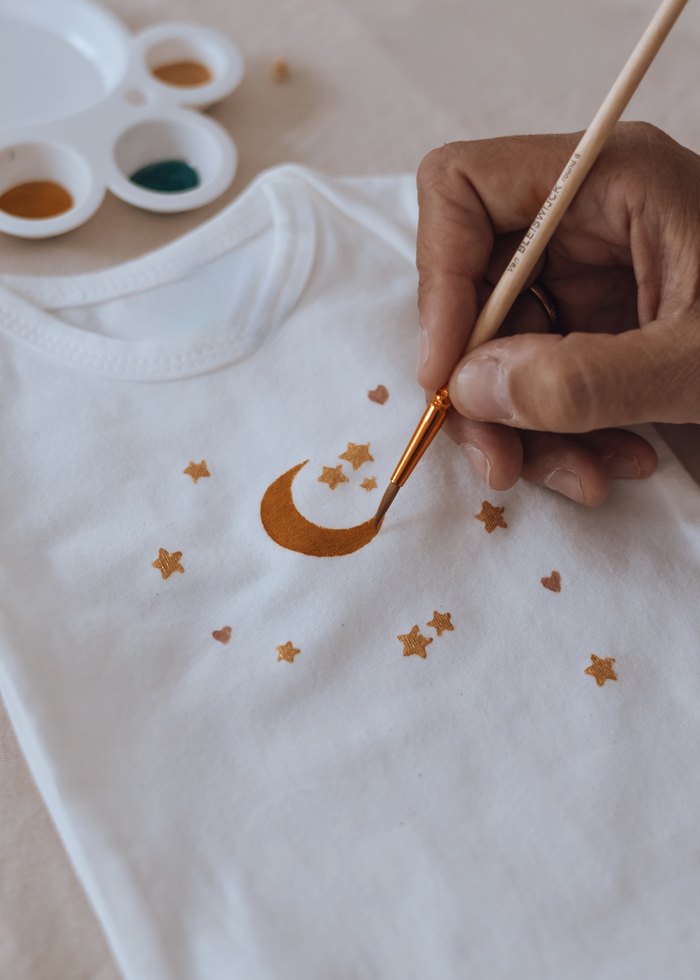 Pinceau doré traçant une lune et des étoiles sur un vêtement de bébé pendant un atelier — Entre Deux, Enghien